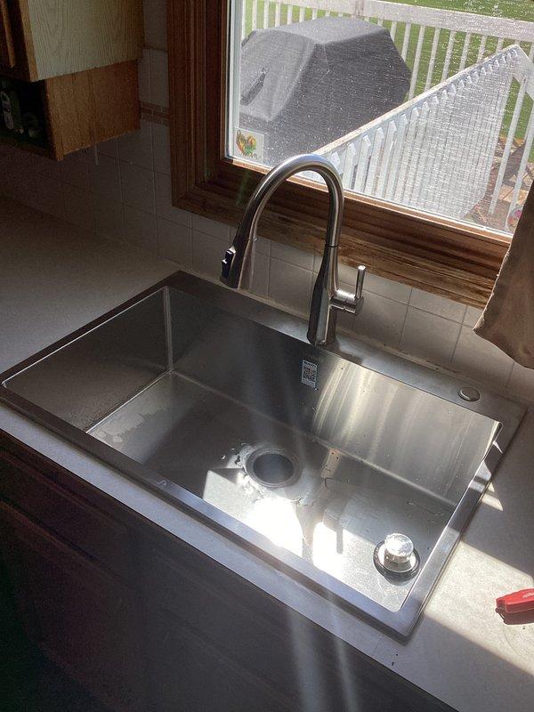 Completed new kitchen sink installation in residential property built in 1997. Installed stainless steel undermount basin with brushed nickel pull-down faucet and integrated garbage disposal unit with red cylindrical body. Connected disposal to drain system using white PVC P-trap configuration, verified all plumbing connections for proper drainage, and completed electrical hookup with standard plug connection to wall outlet. All fixtures tested and operational upon completion.