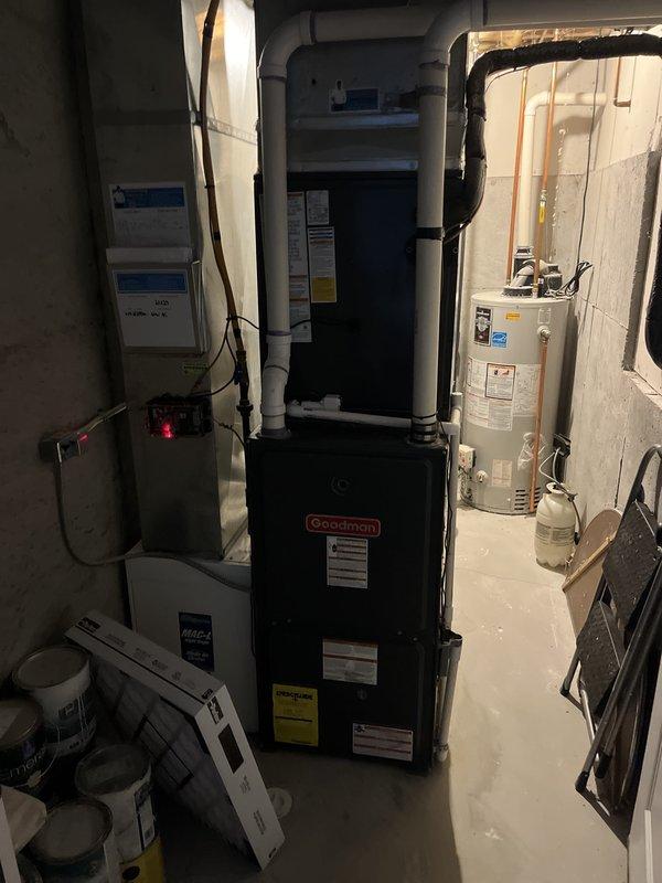 Completed full HVAC system installation including outdoor condensing unit and indoor air handler. Outdoor unit installed on composite pad with proper electrical conduit routing, PVC condensate drain line, and refrigerant line connections. Indoor Goodman air handler installed in basement mechanical room with integrated ductwork connections, electrical service, and proper clearances maintained around existing water heater and utility equipment.