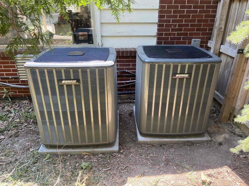 Completed installation of two new outdoor air conditioning condensing units positioned side by side on concrete pads in residential side yard area. Units mounted against building exterior with proper clearances maintained between house wall and perimeter fence. Refrigerant lines and electrical conduit professionally routed along exterior wall to connect both systems. All connections secured, tested for proper operation, and system startup procedures completed successfully.