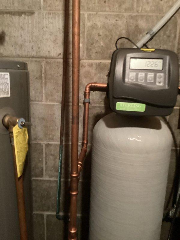 Completed water softener system replacement in residential basement utility area. Removed existing aged water softener unit showing signs of corrosion and wear, then installed new white cylindrical water softener tank with digital control head unit featuring programmable display and multi-button interface. Connected unit to existing copper supply lines using professional soldered joints and appropriate fittings. Verified all plumbing connections for proper flow and tested system operation to ensure digital display and control functions working correctly. Disposed of old equipment per standard procedures.