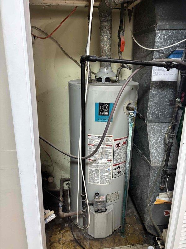 Responded to no cooling call on five-year-old residential air conditioning system affecting entire home. Upon arrival, performed diagnostic assessment of HVAC equipment located in utility closet adjacent to water heater. Inspected air handler unit and associated ductwork in basement area, noting significant deterioration of metal ductwork with visible corrosion and flaking surface finish on 90-degree elbow joint and connected sections. Evaluated mechanical connections, electrical supply, and refrigerant lines to air handler. Documented equipment condition through photographs showing HVAC unit positioning, plumbing connections, and compromised ductwork requiring attention for proper system airflow and efficiency.