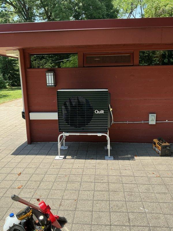 Completed full installation of Quilt brand ductless mini-split system at residential property. Outdoor condenser unit mounted on white metal brackets against exterior wall using concrete pad foundation with metal support posts, with white PVC refrigerant line set and condensate drain properly secured to building siding. System tested and verified operational upon completion of same-day installation.