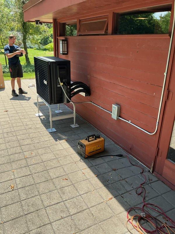 Completed full installation of Quilt brand ductless mini-split system at residential property. Outdoor condenser unit mounted on white metal brackets against exterior wall using concrete pad foundation with metal support posts, with white PVC refrigerant line set and condensate drain properly secured to building siding. System tested and verified operational upon completion of same-day installation.