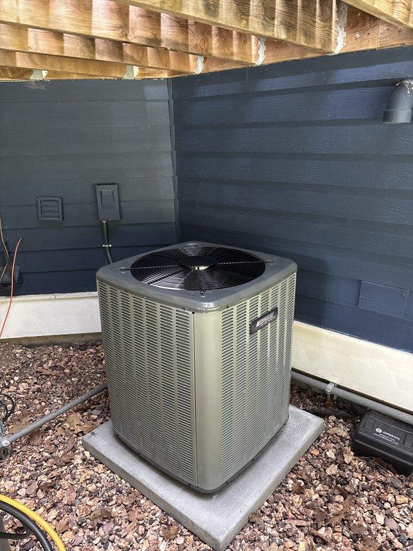 Completed full HVAC system replacement including outdoor condensing unit and indoor air handler. Installed new beige cylindrical condensing unit on existing concrete pad with proper clearances beneath structural joists, positioned against building exterior with wood chip and gravel drainage surrounding foundation. Mounted new dark-colored air handler in interior utility closet with horizontal supply ductwork connection through wall. Verified all electrical connections, refrigerant lines, and ductwork transitions properly secured and sealed for optimal system performance.