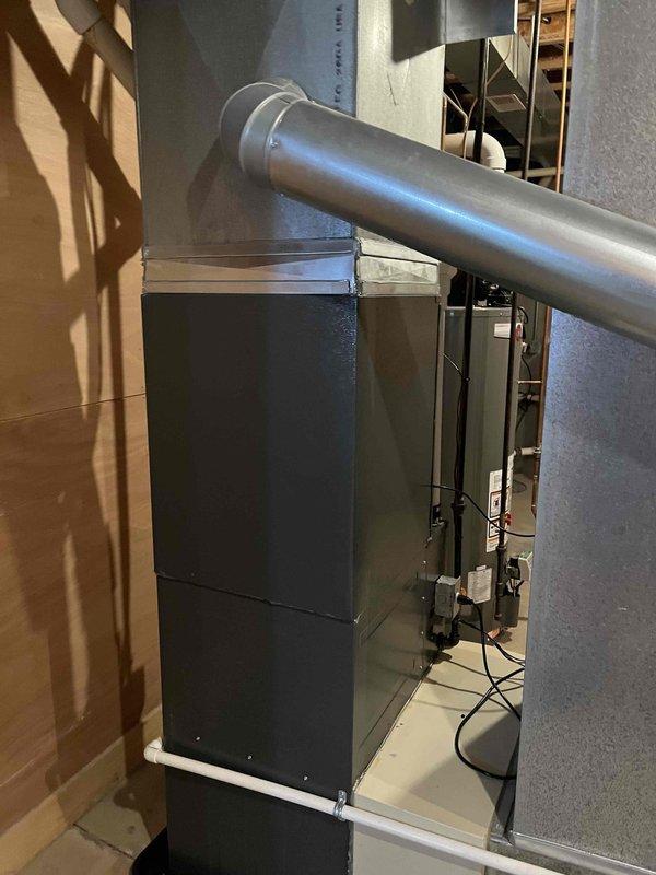 Completed full HVAC system replacement including outdoor condensing unit and indoor air handler. Installed new beige cylindrical condensing unit on existing concrete pad with proper clearances beneath structural joists, positioned against building exterior with wood chip and gravel drainage surrounding foundation. Mounted new dark-colored air handler in interior utility closet with horizontal supply ductwork connection through wall. Verified all electrical connections, refrigerant lines, and ductwork transitions properly secured and sealed for optimal system performance.