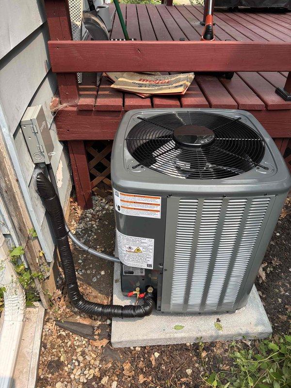 Completed full installation of residential furnace and air conditioning system. New outdoor condensing unit installed on concrete pad adjacent to building exterior beneath existing deck structure, with proper clearances maintained and electrical conduit routed from building wall. System commissioned, tested for proper operation, and documentation photographed for record.