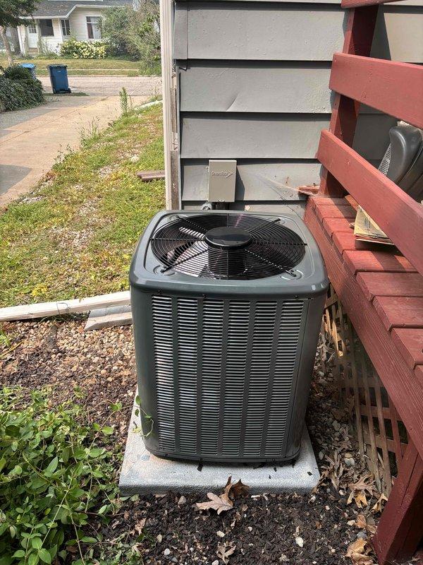 Completed full installation of residential furnace and air conditioning system. New outdoor condensing unit installed on concrete pad adjacent to building exterior beneath existing deck structure, with proper clearances maintained and electrical conduit routed from building wall. System commissioned, tested for proper operation, and documentation photographed for record.