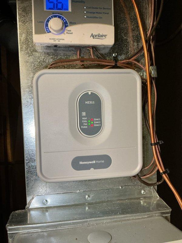 Responded to service call for two-zone air conditioning system with one zone not functioning properly. Upon arrival, inspected 25-year-old system that had been previously upgraded with Honeywell Home HZ311 zone control panel. Evaluated zone damper operation and control wiring connections at equipment location. Examined zone damper actuator motor with red adjustment dial, verified proper mounting and mechanical operation. Tested zone control panel functionality, confirmed digital display and zone indicator lights operational. Checked damper motor positioning and response to thermostat calls. Verified Aprilaire humidifier controller settings and operation as part of overall system assessment. Inspected all visible wiring connections between zone control panel and damper actuators. System evaluation completed with photographic documentation of control equipment and damper actuator installation for service records.