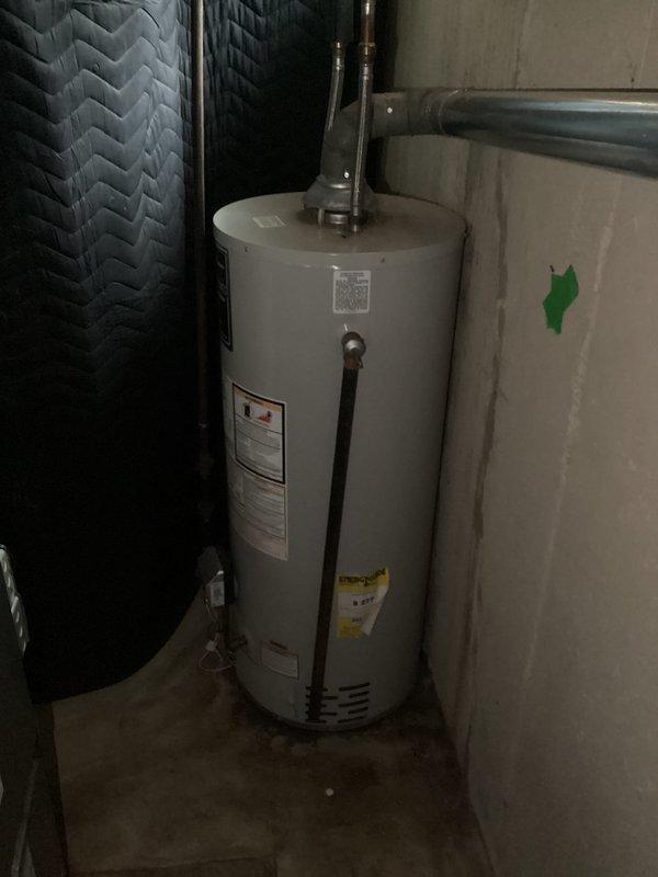 Responded to customer report of leaking water heater and performed comprehensive assessment of existing residential water heater system located in basement utility area. Upon inspection, observed active water staining and discoloration on concrete floor surrounding base of water heater tank, confirming reported leak condition. Documented current installation with beige cylindrical tank, copper supply piping with compression fittings at top connections, and adjacent water treatment equipment. Evaluated plumbing penetrations through walls including galvanized steel piping with rough patching material requiring attention. Photographed equipment condition, leak evidence, and installation configuration to provide accurate estimate for water heater replacement and associated plumbing modifications needed to address the leak and bring installation up to current code standards.