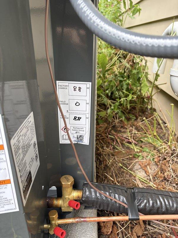Completed full air conditioning system replacement at residential property. Removed existing outdoor condensing unit and installed new equipment on existing concrete pad against exterior wall with beige horizontal siding. Made all necessary electrical connections via conduit from wall-mounted disconnect box to unit. Connected refrigerant lines using copper piping with brass fittings and service valves, secured with red protective caps. Installed flexible conduit for line set protection. Verified factory charge specifications and completed field connections per manufacturer requirements. Ensured proper PVC condensate drain line routing along building exterior. Positioned unit for optimal clearance and serviceability. All electrical, refrigerant, and drainage connections tested and verified operational. System commissioned and left in full working order.