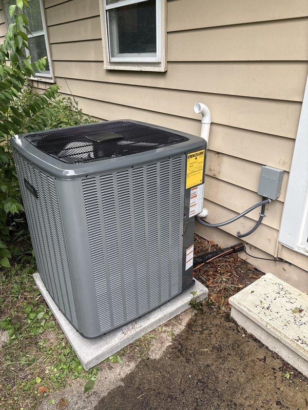 Completed full air conditioning system replacement at residential property. Removed existing outdoor condensing unit and installed new equipment on existing concrete pad against exterior wall with beige horizontal siding. Made all necessary electrical connections via conduit from wall-mounted disconnect box to unit. Connected refrigerant lines using copper piping with brass fittings and service valves, secured with red protective caps. Installed flexible conduit for line set protection. Verified factory charge specifications and completed field connections per manufacturer requirements. Ensured proper PVC condensate drain line routing along building exterior. Positioned unit for optimal clearance and serviceability. All electrical, refrigerant, and drainage connections tested and verified operational. System commissioned and left in full working order.