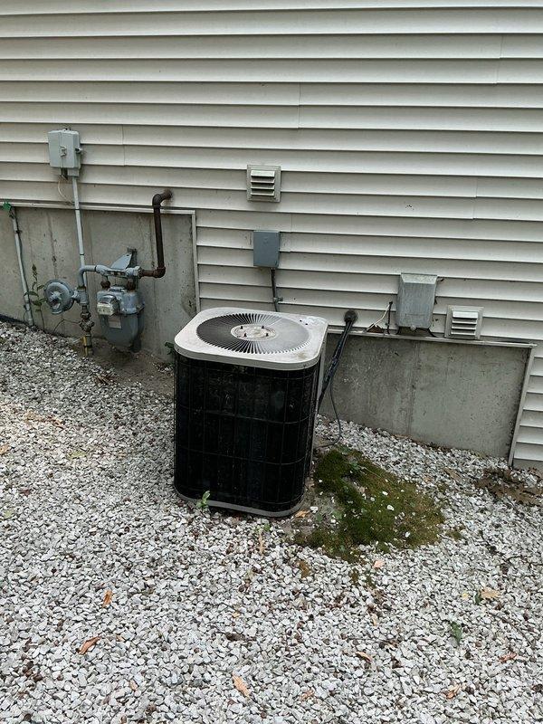 Completed comprehensive sales estimate for furnace and air conditioning system replacement. Existing equipment remains operational but inspection revealed failed heat exchanger in furnace creating hazardous condensation issues in basement area. Exterior AC condenser unit showing significant age-related deterioration with weathering and dirt accumulation on black cylindrical housing. Documented current system configuration including outdoor condensing unit positioned on gravel bed alongside building foundation, utility connections, and basement mechanical room conditions. Assessed unfinished basement space with exposed concrete block walls, wooden ceiling joists, existing ductwork, and electrical infrastructure. Photographed equipment locations, mounting surfaces, and utility routing to support replacement proposal. Provided customer with detailed estimate for new high-efficiency furnace and air conditioning system installation to address failed heat exchanger safety concern and improve