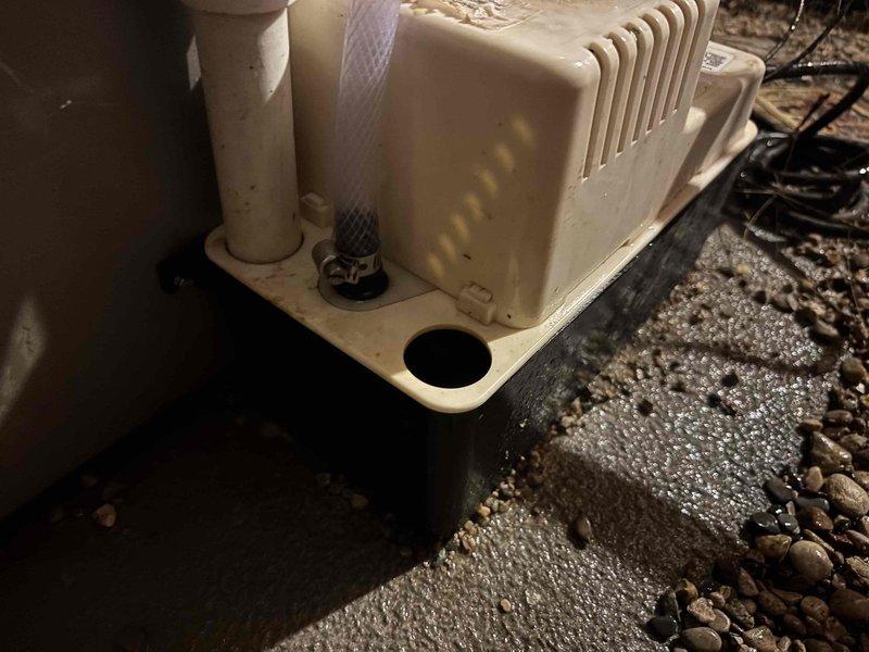 # HVAC Condensate Pump System Service

Responded to a no cooling call and inspected the HVAC condensate management system. Found the condensate pump unit located in a crawl space area with proper drainage pan installation. The cream-colored drain pan showed evidence of water accumulation and mineral deposits on the surrounding concrete surface, indicating the system had been processing condensate but required attention.

Examined the PVC drainage piping configuration, which featured multiple elbows and vertical runs connecting the pump to the overhead discharge point. Verified proper pump operation and drainage flow through the white PVC piping system. The pump unit displayed normal ventilation through its slotted housing design. Confirmed all pipe connections were secure and the drainage system was functioning as designed to remove condensate from the air conditioning unit effectively.