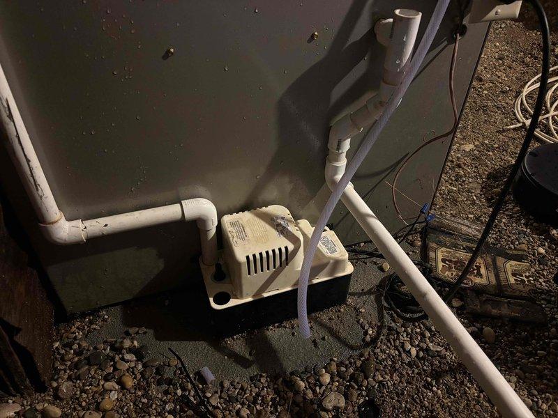 # HVAC Condensate Pump System Service

Responded to a no cooling call and inspected the HVAC condensate management system. Found the condensate pump unit located in a crawl space area with proper drainage pan installation. The cream-colored drain pan showed evidence of water accumulation and mineral deposits on the surrounding concrete surface, indicating the system had been processing condensate but required attention.

Examined the PVC drainage piping configuration, which featured multiple elbows and vertical runs connecting the pump to the overhead discharge point. Verified proper pump operation and drainage flow through the white PVC piping system. The pump unit displayed normal ventilation through its slotted housing design. Confirmed all pipe connections were secure and the drainage system was functioning as designed to remove condensate from the air conditioning unit effectively.