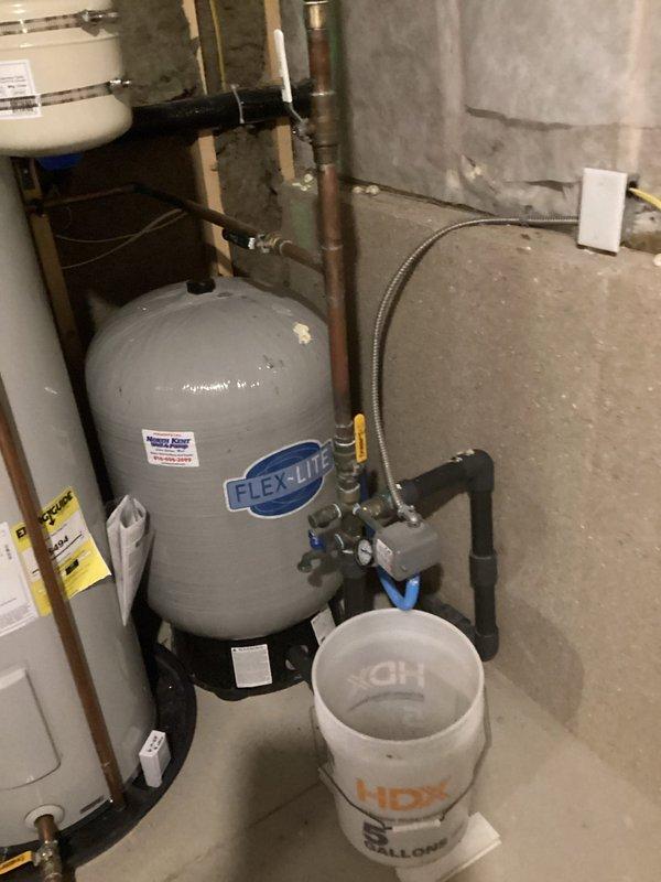 Responded to plumbing leak complaint regarding rusting well pressure tank in basement. Upon inspection, found gray FLEXLITE pressure tank showing signs of corrosion and active leaking, consistent with hard water conditions prevalent in the area. Documented existing tank condition with copper and black pipe connections, blue-handled shutoff valves, and flexible braided hoses. Noted mechanical assembly with yellow-handled control valve and pressure gauge mounted on concrete block wall. Placed collection bucket under leak point to prevent further water damage to basement floor. Tank deterioration attributed to aggressive hard water chemistry causing accelerated rust-through of tank shell, a common failure mode for well pressure tanks in this region. Photographed complete installation including plumbing connections, valve configuration, and current leak conditions for service records and potential replacement planning.