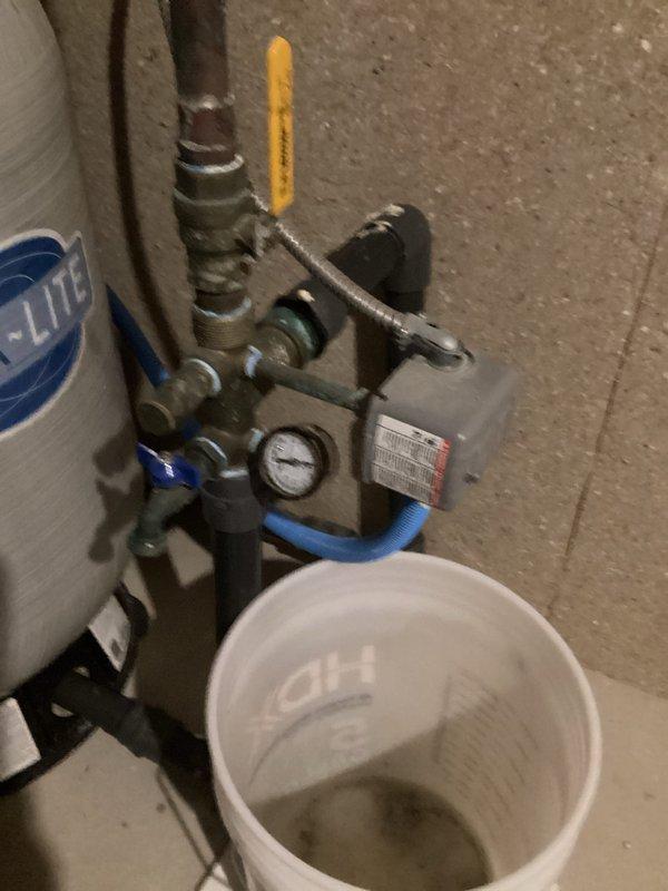Responded to plumbing leak complaint regarding rusting well pressure tank in basement. Upon inspection, found gray FLEXLITE pressure tank showing signs of corrosion and active leaking, consistent with hard water conditions prevalent in the area. Documented existing tank condition with copper and black pipe connections, blue-handled shutoff valves, and flexible braided hoses. Noted mechanical assembly with yellow-handled control valve and pressure gauge mounted on concrete block wall. Placed collection bucket under leak point to prevent further water damage to basement floor. Tank deterioration attributed to aggressive hard water chemistry causing accelerated rust-through of tank shell, a common failure mode for well pressure tanks in this region. Photographed complete installation including plumbing connections, valve configuration, and current leak conditions for service records and potential replacement planning.