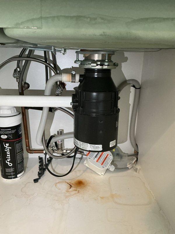 Responded to kitchen sink leak in 1991 single-family home where water was actively leaking onto the kitchen floor from under the sink cabinet. Upon arrival, performed thorough inspection of all plumbing components beneath the kitchen sink, including garbage disposal unit, drain connections, water supply lines, copper piping, flexible supply hoses, shutoff valves, PVC drain system, and water filtration system. Identified source of leak and completed necessary repairs to stop water infiltration. Verified all connections at disposal unit mounting flange, corrugated drain hoses with metal hose clamps, and braided supply lines were properly secured. Tested system under full water pressure to confirm leak was resolved and no additional moisture was present. Documented existing water stains on cabinet floor from previous leak occurrence. All plumbing fixtures returned to proper working condition with no further leakage detected.