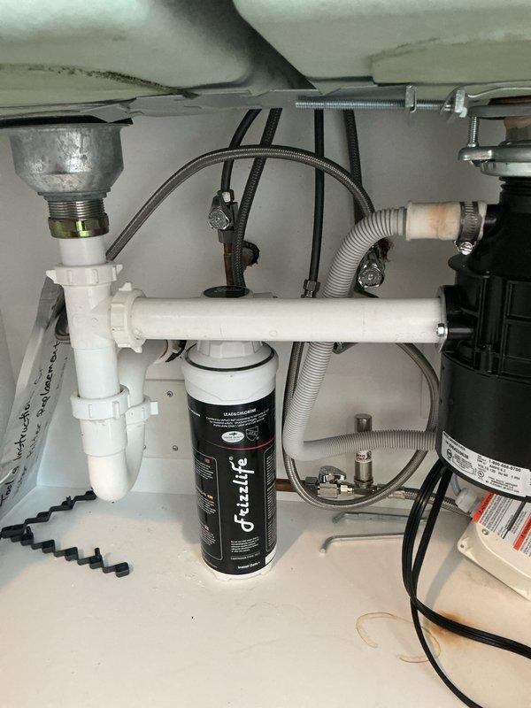 Responded to kitchen sink leak in 1991 single-family home where water was actively leaking onto the kitchen floor from under the sink cabinet. Upon arrival, performed thorough inspection of all plumbing components beneath the kitchen sink, including garbage disposal unit, drain connections, water supply lines, copper piping, flexible supply hoses, shutoff valves, PVC drain system, and water filtration system. Identified source of leak and completed necessary repairs to stop water infiltration. Verified all connections at disposal unit mounting flange, corrugated drain hoses with metal hose clamps, and braided supply lines were properly secured. Tested system under full water pressure to confirm leak was resolved and no additional moisture was present. Documented existing water stains on cabinet floor from previous leak occurrence. All plumbing fixtures returned to proper working condition with no further leakage detected.