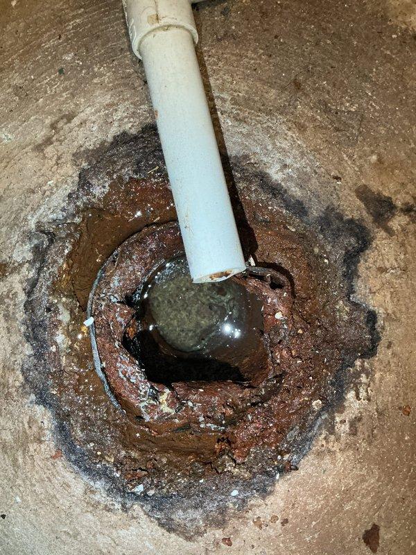 Responded to a plumbing service call for a floor drain experiencing slow drainage, with the drain serving only the furnace, air conditioner, and humidifier condensate lines. Upon inspection, discovered a heavily corroded metal floor drain fitting embedded in the concrete floor with extensive rust deterioration, pitting, and brown-orange discoloration throughout the metal components. The drain opening showed significant age-related degradation with rust staining radiating outward onto the surrounding concrete surface. A white PVC pipe connected vertically to the corroded drain fitting, serving as the condensate discharge line from the HVAC equipment. The metal drain exhibited rough, pitted texture indicating advanced corrosion that was likely contributing to reduced drainage capacity and flow restriction. Documented the condition of the corroded floor drain assembly, the PVC condensate line connection, and the extent of rust damage affecting the drain performance. The deteriorated state