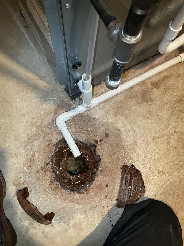Responded to a plumbing service call for a floor drain experiencing slow drainage, with the drain serving only the furnace, air conditioner, and humidifier condensate lines. Upon inspection, discovered a heavily corroded metal floor drain fitting embedded in the concrete floor with extensive rust deterioration, pitting, and brown-orange discoloration throughout the metal components. The drain opening showed significant age-related degradation with rust staining radiating outward onto the surrounding concrete surface. A white PVC pipe connected vertically to the corroded drain fitting, serving as the condensate discharge line from the HVAC equipment. The metal drain exhibited rough, pitted texture indicating advanced corrosion that was likely contributing to reduced drainage capacity and flow restriction. Documented the condition of the corroded floor drain assembly, the PVC condensate line connection, and the extent of rust damage affecting the drain performance. The deteriorated state