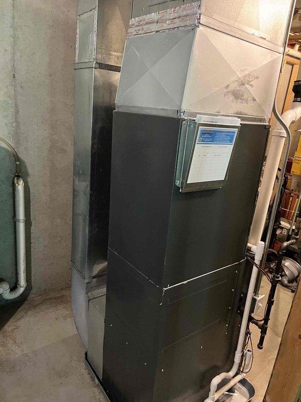 Complete HVAC system replacement performed, including installation of new air handler unit in basement utility area and outdoor condensing unit. Air handler positioned against concrete wall with proper ductwork connections established at ceiling level and pyramidal cap installed for optimal airflow distribution. Unit secured with all technical documentation affixed to front panel for easy reference. Outdoor condensing unit installed on concrete pad against exterior wall with beige vinyl siding, positioned adjacent to stacked concrete block retaining wall. Unit placement optimized for accessibility while maintaining aesthetic integration with existing landscaping. All refrigerant lines connected between indoor and outdoor components, electrical connections completed to manufacturer specifications, and system tested for proper operation. Condensate drainage verified and all safety controls confirmed functional. System startup completed with airflow balanced and thermostat communication e