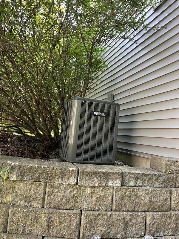 Complete HVAC system replacement performed, including installation of new air handler unit in basement utility area and outdoor condensing unit. Air handler positioned against concrete wall with proper ductwork connections established at ceiling level and pyramidal cap installed for optimal airflow distribution. Unit secured with all technical documentation affixed to front panel for easy reference. Outdoor condensing unit installed on concrete pad against exterior wall with beige vinyl siding, positioned adjacent to stacked concrete block retaining wall. Unit placement optimized for accessibility while maintaining aesthetic integration with existing landscaping. All refrigerant lines connected between indoor and outdoor components, electrical connections completed to manufacturer specifications, and system tested for proper operation. Condensate drainage verified and all safety controls confirmed functional. System startup completed with airflow balanced and thermostat communication e