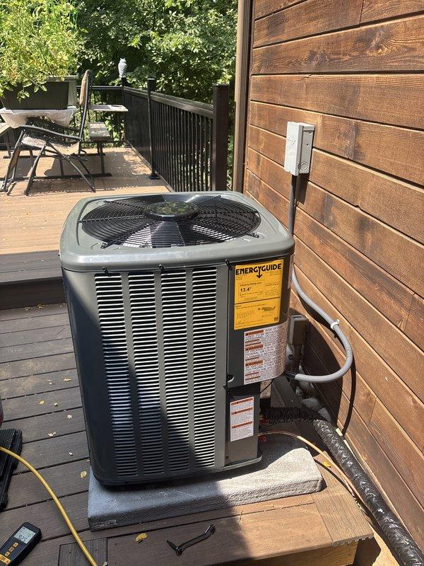 Successfully completed full air conditioning system replacement at residential property. Removed existing outdoor condensing unit and installed new high-efficiency AC condenser on wooden deck adjacent to exterior wall. Unit positioned on protective rubber pad with proper clearances maintained for airflow and serviceability.

Installed new refrigerant lines with black insulation running from outdoor unit through exterior wall to indoor air handler. Mounted electrical disconnect box on exterior siding per code requirements. System charged with refrigerant per manufacturer specifications - factory charge of 54 oz plus field charge of 26 oz for total system charge of 80 oz. Refrigerant type classified as A2L safety group per updated environmental standards.

Connected and secured all copper refrigerant lines with brass service valves and protective caps installed on both suction and liquid lines. Verified all electrical connections, tested unit operation including compressor and condenser 