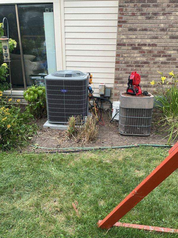 Completed full residential air conditioning system replacement including outdoor condenser unit and indoor air handler components. Removed existing aged cylindrical condenser unit showing significant weathering and corrosion, and installed new gray rectangular high-efficiency condensing unit on exterior concrete pad against brick foundation. Positioned new equipment in existing location adjacent to house with proper clearances for airflow and service access. 

Installed new evaporator coil and air handler in basement mechanical room, integrating with existing furnace system. Ran refrigerant line set between outdoor condenser and indoor coil through building envelope, ensuring proper insulation and weatherproofing of penetrations. Connected white PVC condensate drain piping from evaporator coil with appropriate trap and routing to existing drain system. Installed electrical disconnect at outdoor unit per code requirements.

Pressure tested refrigerant lines for leaks, evacuated system t