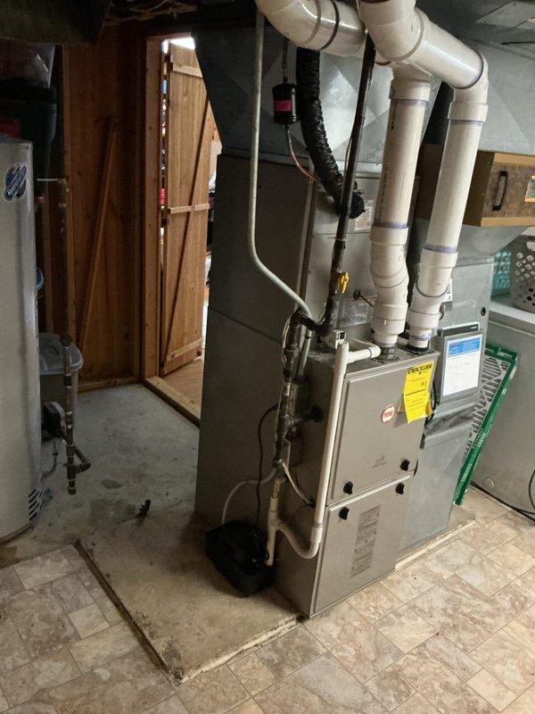 Completed full residential air conditioning system replacement including outdoor condenser unit and indoor air handler components. Removed existing aged cylindrical condenser unit showing significant weathering and corrosion, and installed new gray rectangular high-efficiency condensing unit on exterior concrete pad against brick foundation. Positioned new equipment in existing location adjacent to house with proper clearances for airflow and service access. 

Installed new evaporator coil and air handler in basement mechanical room, integrating with existing furnace system. Ran refrigerant line set between outdoor condenser and indoor coil through building envelope, ensuring proper insulation and weatherproofing of penetrations. Connected white PVC condensate drain piping from evaporator coil with appropriate trap and routing to existing drain system. Installed electrical disconnect at outdoor unit per code requirements.

Pressure tested refrigerant lines for leaks, evacuated system t