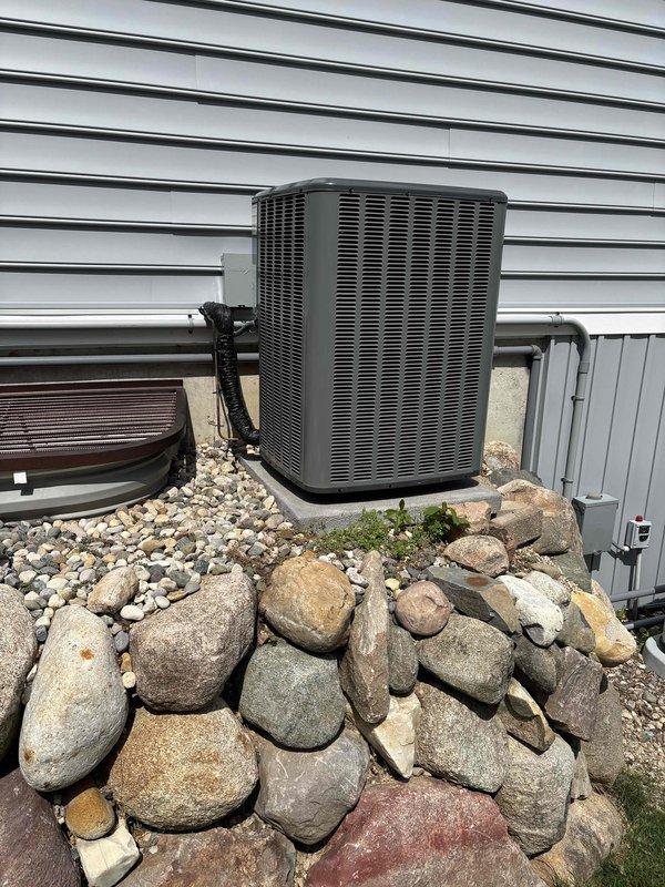 Complete HVAC system installation performed, including new furnace and air conditioning condenser unit. Furnace installed in basement utility area with proper PVC venting routed vertically through ceiling, integrated with existing ductwork system using metallic and flexible ducting. AC condenser unit positioned on exterior rock retaining wall against building with proper refrigerant line connections, electrical disconnect box mounted to siding, and all required electrical conduit installed. All equipment secured, tested, and verified operational per manufacturer specifications.