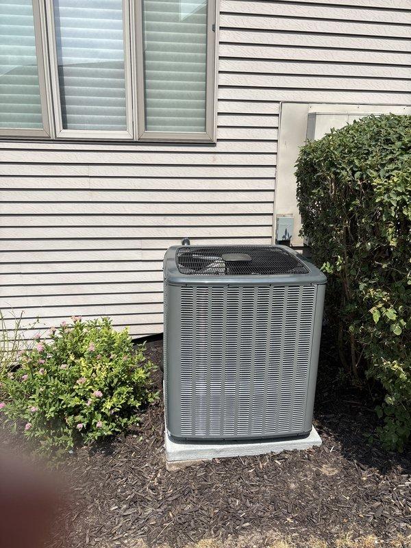 Completed full air conditioning system replacement at residential property. Removed existing outdoor condensing unit and installed new replacement unit on concrete pad against house exterior with white vinyl siding. New unit positioned in landscaped area with mulch bedding and surrounding shrubbery. Installed copper refrigerant line set with proper insulation and brass fittings, ensuring all connections were secure and leak-free. Verified electrical specifications and proper unit operation. System tested and commissioned with performance parameters documented, including temperature differential readings displayed on service tag. Installation completed per manufacturer specifications with all safety and code requirements met.
