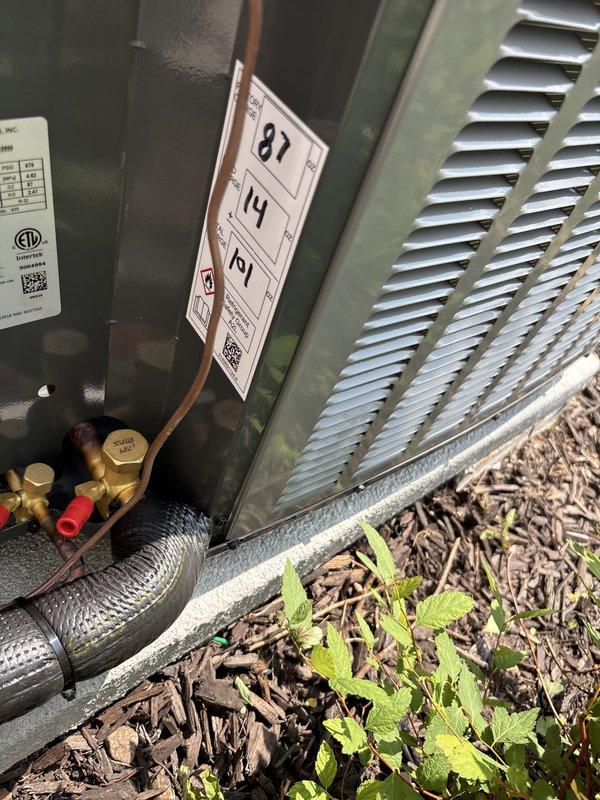 Completed full air conditioning system replacement at residential property. Removed existing outdoor condensing unit and installed new replacement unit on concrete pad against house exterior with white vinyl siding. New unit positioned in landscaped area with mulch bedding and surrounding shrubbery. Installed copper refrigerant line set with proper insulation and brass fittings, ensuring all connections were secure and leak-free. Verified electrical specifications and proper unit operation. System tested and commissioned with performance parameters documented, including temperature differential readings displayed on service tag. Installation completed per manufacturer specifications with all safety and code requirements met.