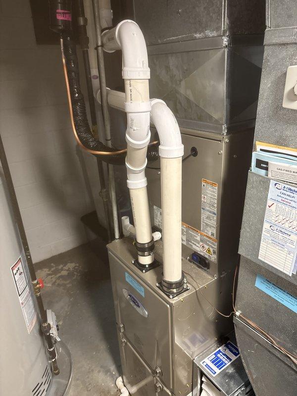 Completed full air conditioning system replacement in basement utility area. Removed existing equipment and installed new gray metal air handler unit with proper mounting and securing to concrete block wall. Ran and secured copper refrigerant lines with black insulation along wall, connecting outdoor condensing unit to indoor air handler. Installed white PVC condensate drain piping with proper pitch and securing, running vertically from air handler with appropriate elbow fittings and connections. Connected all electrical wiring to new equipment per manufacturer specifications. Integrated new system with existing ductwork, ensuring proper sealing at all connection points. Verified all refrigerant line connections were properly brazed and pressure tested. Confirmed condensate drainage system functioning correctly. Started up system, checked refrigerant charge, verified proper airflow through ductwork, and tested all operational modes. System operating within manufacturer specifications w