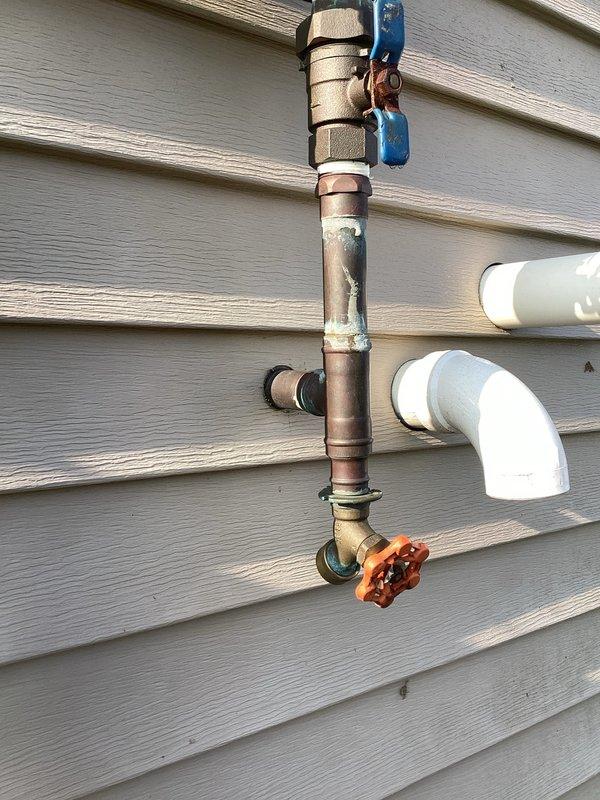 Responded to outdoor irrigation spigot with chronic leak that had progressively worsened over three years from minor drip to constant flow. Upon inspection, found aged copper piping assembly with oxidation and weathering on 21-year-old residential exterior plumbing system. Evaluated vertical copper pipe configuration with backflow preventer, multiple brass fittings, compression couplings, and gate valve with wheel handle. Assessed condition of existing shutoff mechanisms including blue-handled valve at top of assembly and brass valve at lower section. Documented deteriorated connections and T-fitting junction points where leak originated. Addressed improperly capped spout that had been temporary solution. Repaired or replaced failed components in irrigation system plumbing to restore proper function and eliminate constant water loss.