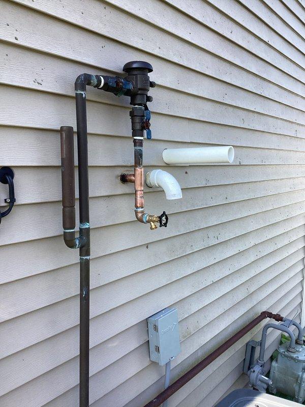 Responded to outdoor irrigation spigot with chronic leak that had progressively worsened over three years from minor drip to constant flow. Upon inspection, found aged copper piping assembly with oxidation and weathering on 21-year-old residential exterior plumbing system. Evaluated vertical copper pipe configuration with backflow preventer, multiple brass fittings, compression couplings, and gate valve with wheel handle. Assessed condition of existing shutoff mechanisms including blue-handled valve at top of assembly and brass valve at lower section. Documented deteriorated connections and T-fitting junction points where leak originated. Addressed improperly capped spout that had been temporary solution. Repaired or replaced failed components in irrigation system plumbing to restore proper function and eliminate constant water loss.