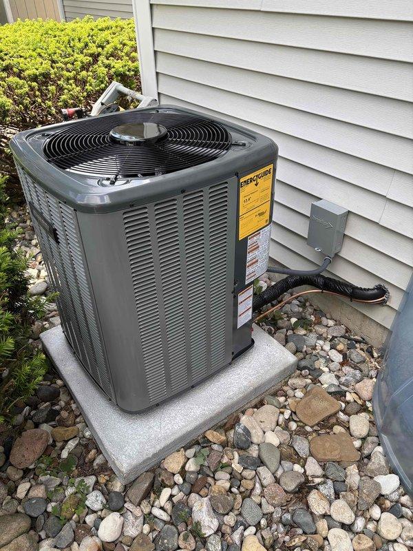 Completed full HVAC system installation including new furnace and air conditioning unit. Indoor furnace unit installed in mechanical room on protective base pad with proper PVC venting routed from top of unit, connected to existing ductwork system with digital thermostat control. Outdoor condensing unit positioned on concrete pad against exterior wall with electrical disconnect box mounted on siding, refrigerant lines and drainage properly installed, unit surrounded by decorative stone landscaping for drainage and aesthetics. All equipment labeled with manufacturer specifications, system tested and operational.