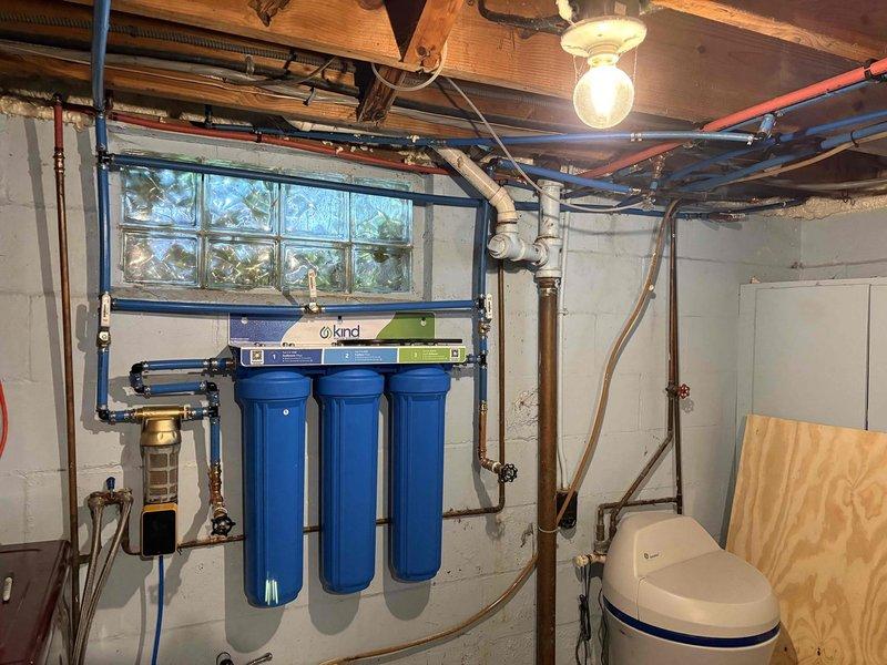 Completed comprehensive repiping of water distribution lines in basement utility area, replacing existing plumbing with new PEX and copper piping infrastructure. Installed customer-supplied three-stage water filtration system with blue cylindrical filter housings and KIND brand control panel, mounting unit securely to cinderblock wall. Configured manifold system with brass fittings and shut-off valves to integrate filtration system with existing water softener tank and main water distribution lines. All connections pressure-tested and verified for proper operation.