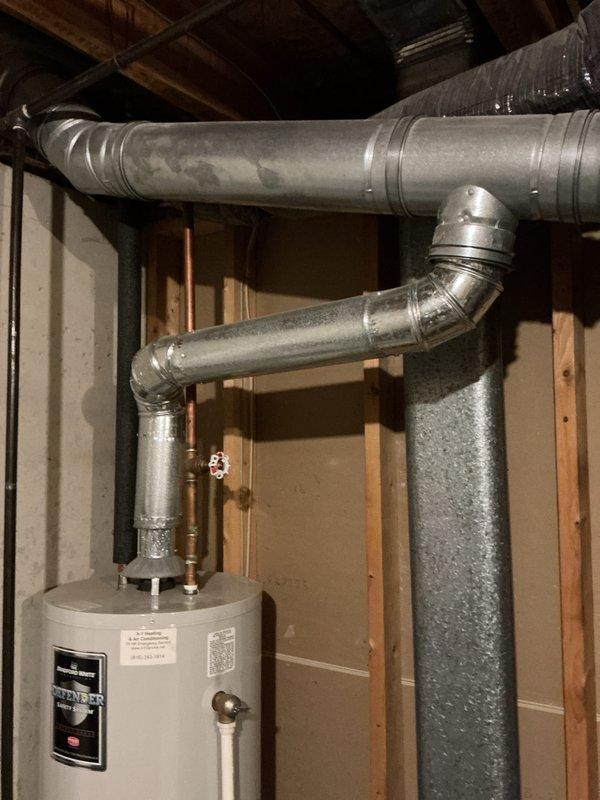 Provided estimate for water heater replacement in basement utility area. Existing white cylindrical water heater assessed with copper piping connections, pressure relief valve, and current integration with overhead HVAC ductwork system. Documented installation space including exposed wooden joists, OSB ceiling panels, galvanized metal ventilation ducts, and structural framing to determine optimal replacement configuration and ensure proper clearances for new unit installation.