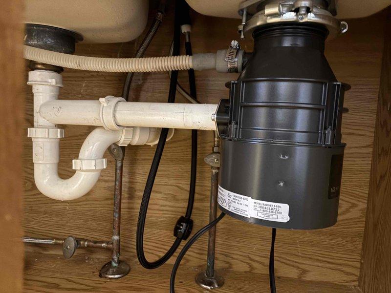 Responded to plumbing leak complaint at kitchen sink location. Upon inspection, identified active leak originating from lower section of garbage disposal unit in double-basin sink configuration. Examined disposal mounting assembly, drain connections, and associated plumbing components including white PVC P-trap system. Documented existing conditions showing black ribbed garbage disposal unit with metal mounting bracket and corrugated discharge hose connection to drain line. Assessed leak source at disposal housing and connection points to determine appropriate repair solution.