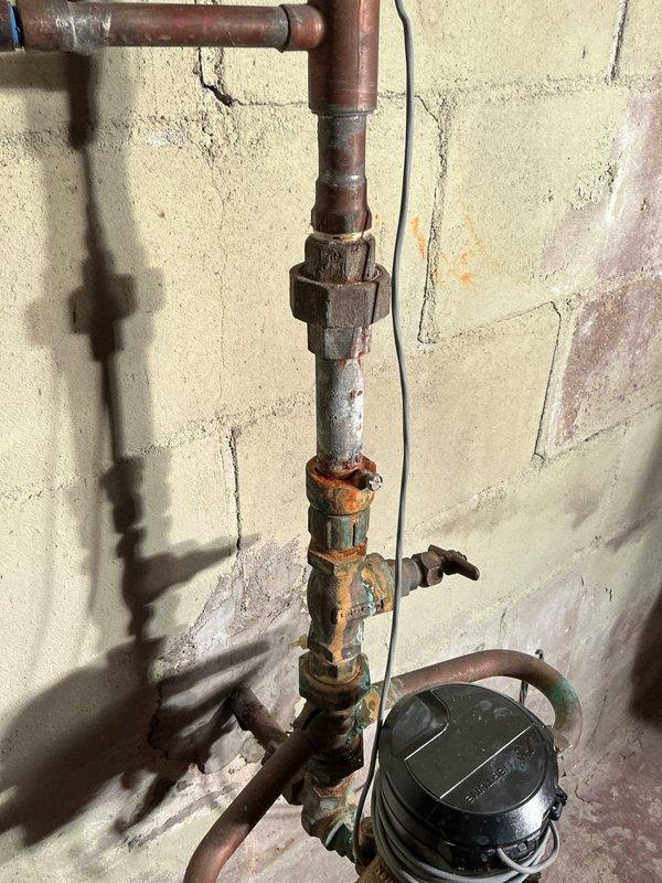 # Plumbing Leak Inspection and Assessment

Responded to service call for water leak originating from main water line at the point where supply lines split to service the building. Upon arrival, performed comprehensive visual inspection of the existing plumbing assembly mounted on exterior wall. Documentation revealed vertical copper piping system with multiple brass fittings, shutoff valves, and water meter installation showing significant age-related deterioration. Observed heavy oxidation and corrosion throughout the copper pipes, with greenish-blue patina formation on brass valve components indicating long-term moisture exposure and material breakdown. Identified multiple compromised connection points along the vertical run where deterioration was most severe. Photographed entire plumbing configuration including horizontal supply line, vertical distribution assembly, brass couplings, valve mechanisms, and meter installation to document current condition and facilitate accurate repla