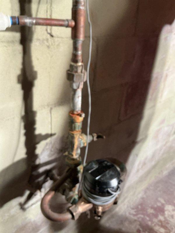 # Plumbing Leak Inspection and Assessment

Responded to service call for water leak originating from main water line at the point where supply lines split to service the building. Upon arrival, performed comprehensive visual inspection of the existing plumbing assembly mounted on exterior wall. Documentation revealed vertical copper piping system with multiple brass fittings, shutoff valves, and water meter installation showing significant age-related deterioration. Observed heavy oxidation and corrosion throughout the copper pipes, with greenish-blue patina formation on brass valve components indicating long-term moisture exposure and material breakdown. Identified multiple compromised connection points along the vertical run where deterioration was most severe. Photographed entire plumbing configuration including horizontal supply line, vertical distribution assembly, brass couplings, valve mechanisms, and meter installation to document current condition and facilitate accurate repla