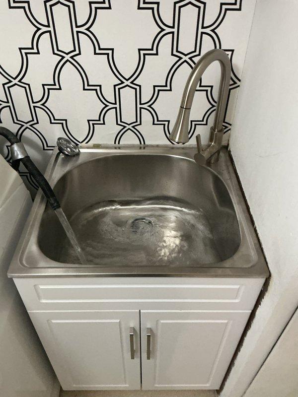 Replaced existing laundry tub with new stainless steel utility sink mounted on white cabinet base with brushed nickel hardware. Installed modern pull-down faucet with brushed nickel finish and connected supply lines. Installed standpipe assembly for washing machine discharge with proper drainage connections and secured all plumbing fixtures to code specifications.