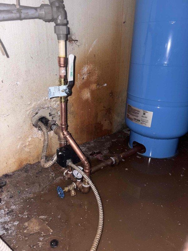 Replaced failed well pressure tank and associated piping in flooded utility area. Removed old tank and deteriorated copper and PVC connections, installed new blue pressure tank with updated piping configuration including flexible connector lines, shut-off valves, and pressure relief devices. Addressed water damage situation and restored proper well water system operation.
