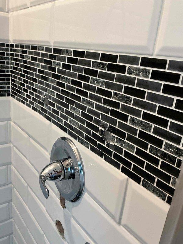 Relocated and installed shower valve assembly in bathroom with decorative tile work. Accessed plumbing through wall cavity, repositioned PEX supply lines and valve body, then mounted chrome shower valve with escutcheon plate on white subway tile section below mosaic glass tile accent band. Secured all connections and restored wall access point.