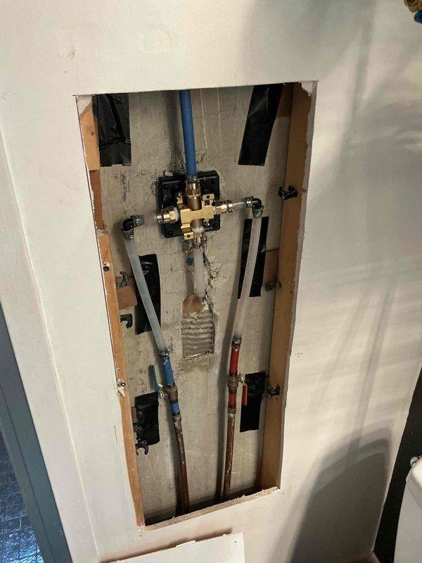 Relocated and installed shower valve assembly in bathroom with decorative tile work. Accessed plumbing through wall cavity, repositioned PEX supply lines and valve body, then mounted chrome shower valve with escutcheon plate on white subway tile section below mosaic glass tile accent band. Secured all connections and restored wall access point.