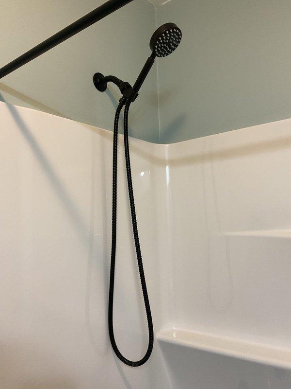 Completed installation of black matte finish shower and tub trim kit in bathroom, including handheld showerhead with wall-mounted bracket, flexible hose, tub spout, faucet handle, and drain controls. All fixtures properly mounted and functional against white shower surround with integrated shelving. Trim kit replacement to be completed on return visit.