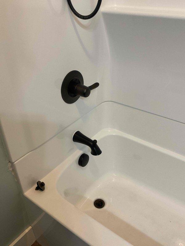 Completed installation of black matte finish shower and tub trim kit in bathroom, including handheld showerhead with wall-mounted bracket, flexible hose, tub spout, faucet handle, and drain controls. All fixtures properly mounted and functional against white shower surround with integrated shelving. Trim kit replacement to be completed on return visit.