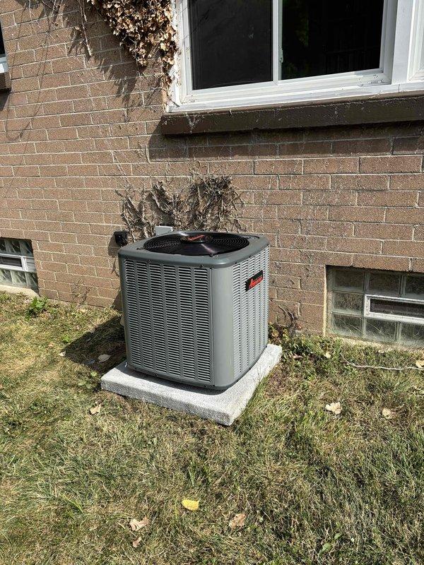 Complete residential air conditioning system replacement performed at single-family home. Removed existing outdoor condensing unit and installed new cylindrical-style condenser unit with horizontal ventilation slats and protective top grille on existing concrete pad adjacent to brick exterior wall. Unit positioned near building corner with proper clearance for airflow and service access. Connected refrigerant lines, electrical supply, and condensate drainage per manufacturer specifications. Verified all utility connections secure and properly routed along exterior wall. System startup completed with successful operational testing of compressor, condenser fan motor, and cooling cycle. New installation features improved energy efficiency and updated safety components including mesh protective grille over fan assembly.