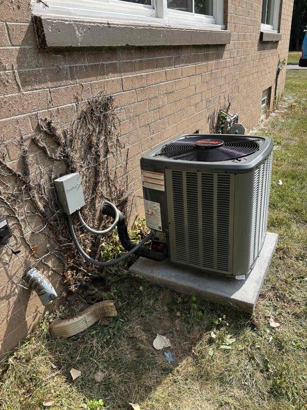 Complete residential air conditioning system replacement performed at single-family home. Removed existing outdoor condensing unit and installed new cylindrical-style condenser unit with horizontal ventilation slats and protective top grille on existing concrete pad adjacent to brick exterior wall. Unit positioned near building corner with proper clearance for airflow and service access. Connected refrigerant lines, electrical supply, and condensate drainage per manufacturer specifications. Verified all utility connections secure and properly routed along exterior wall. System startup completed with successful operational testing of compressor, condenser fan motor, and cooling cycle. New installation features improved energy efficiency and updated safety components including mesh protective grille over fan assembly.