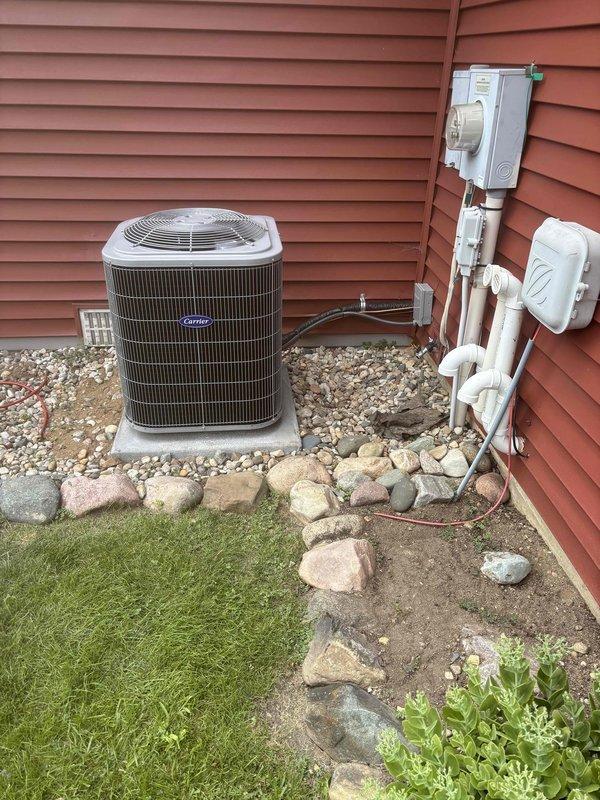Completed full HVAC system installation including new Carrier air conditioning unit and furnace. Outdoor AC unit mounted on concrete pad with proper gravel bedding and decorative rock border, electrical disconnect box installed with copper refrigerant lines and conduit properly routed along exterior wall. Indoor furnace installed in basement utility area with metallic vent piping, integrated humidifier unit with copper connections, and all necessary ductwork connections completed. System tested and confirmed operational.
